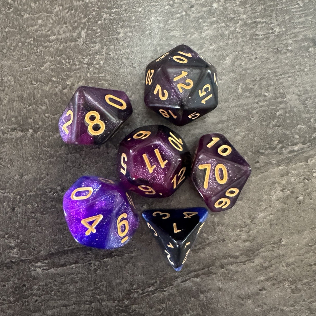 Lot de 6 dés polyédriques violets et sombres pailletés, chiffres dorés. Photo réelle recadrée. / Assortment of 6 purple and dark glitter polyhedral dice with gold-coloured numbers.