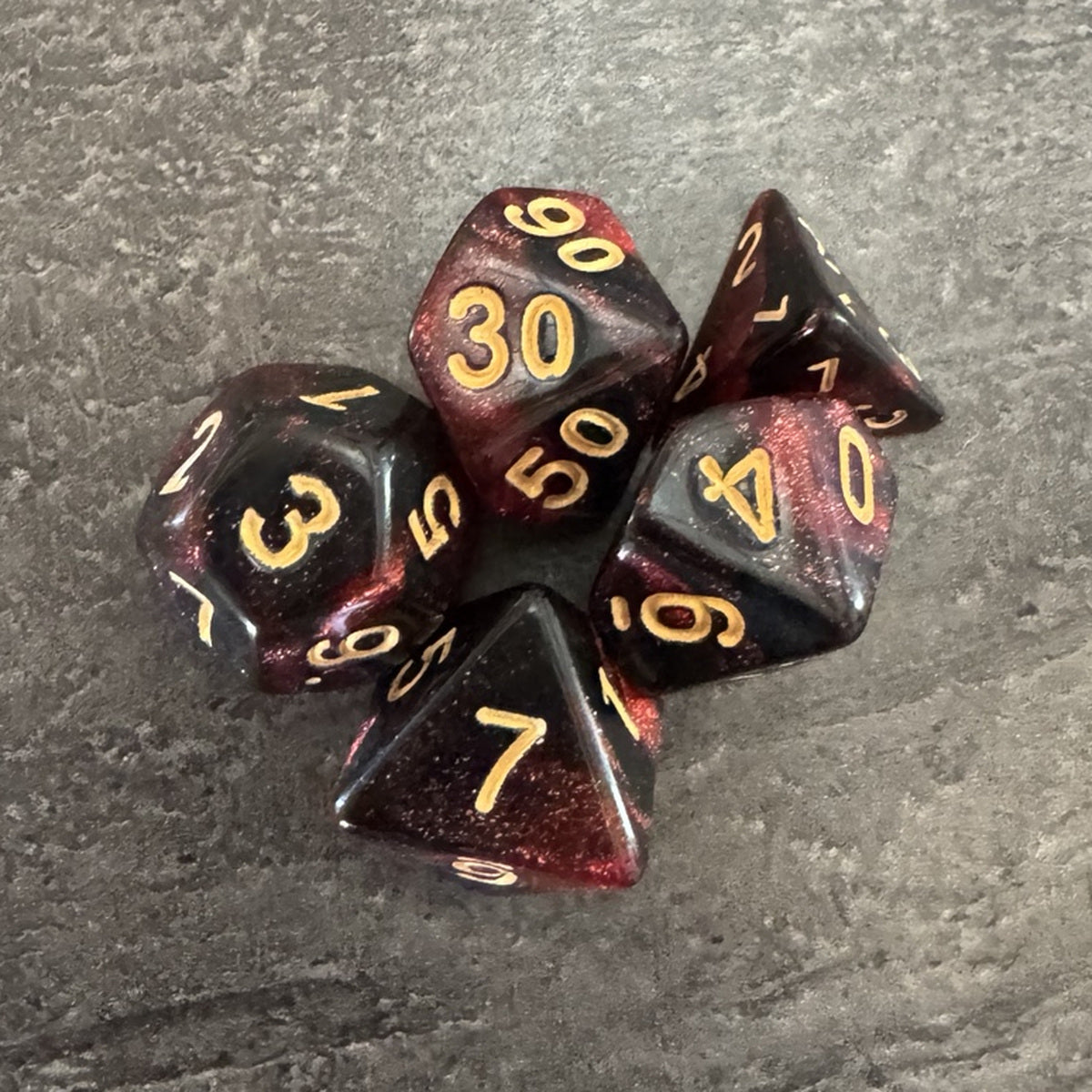 Lot de 5 dés polyédriques rouge et noir pailletés, chiffres dorés. Photo réelle recadrée. / Assortment of 5 red and black glitter polyhedral dice with gold-coloured numbers.