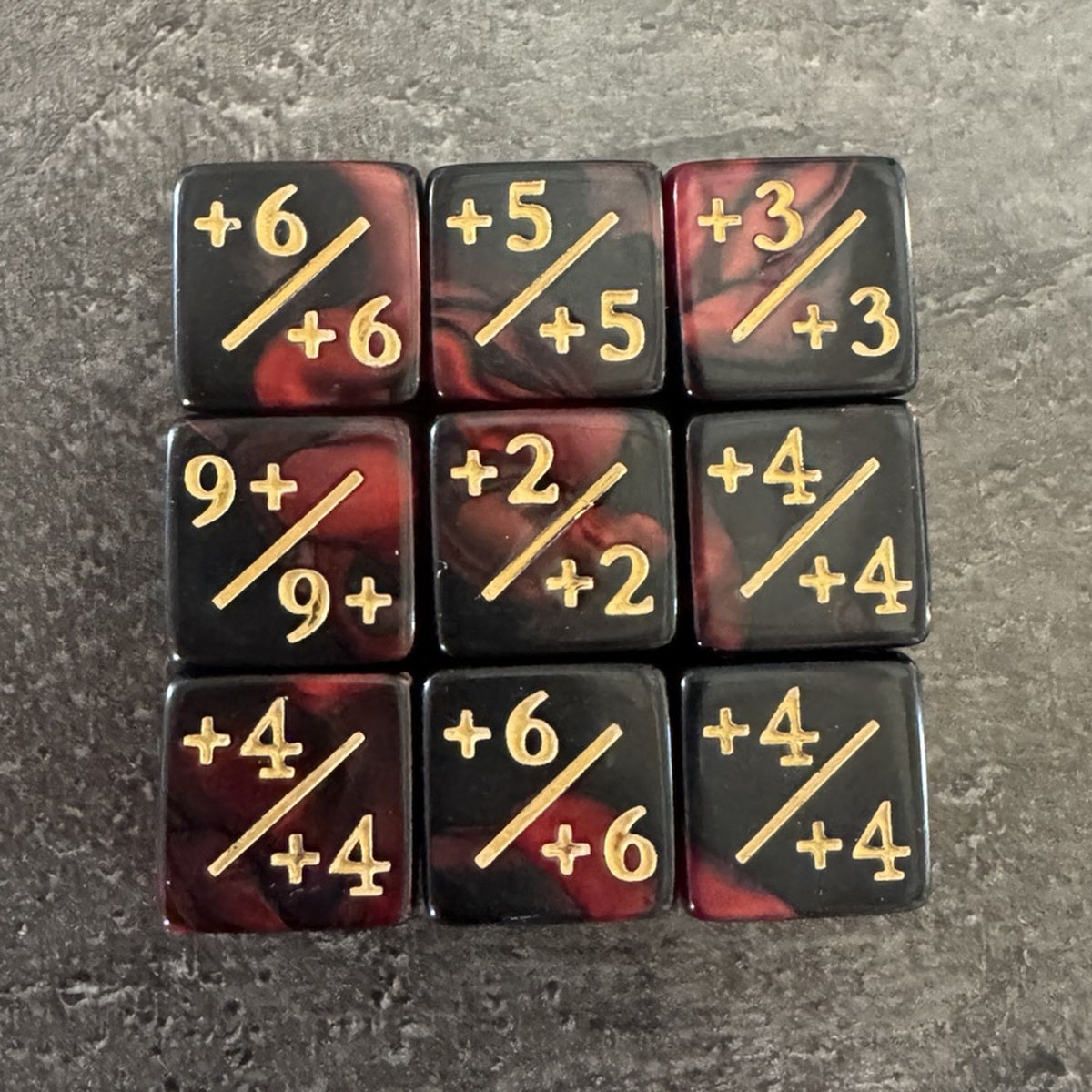 Photo réelle recadrée : 9 dés compteurs positifs rouge et noir, inscriptions dorées +X/+X. / Cropped original photo: 9 red and black positive counter dice.