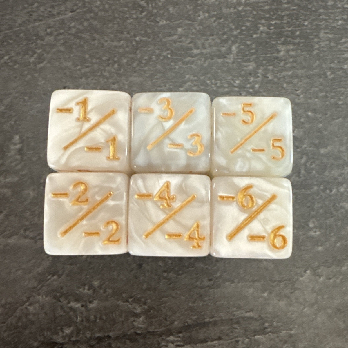 Photo réelle recadrée : 6 dés compteurs négatifs blanc nacré et doré. / Cropped original photo: 6 pearlescent white negative counter dice with gold-coloured markings.