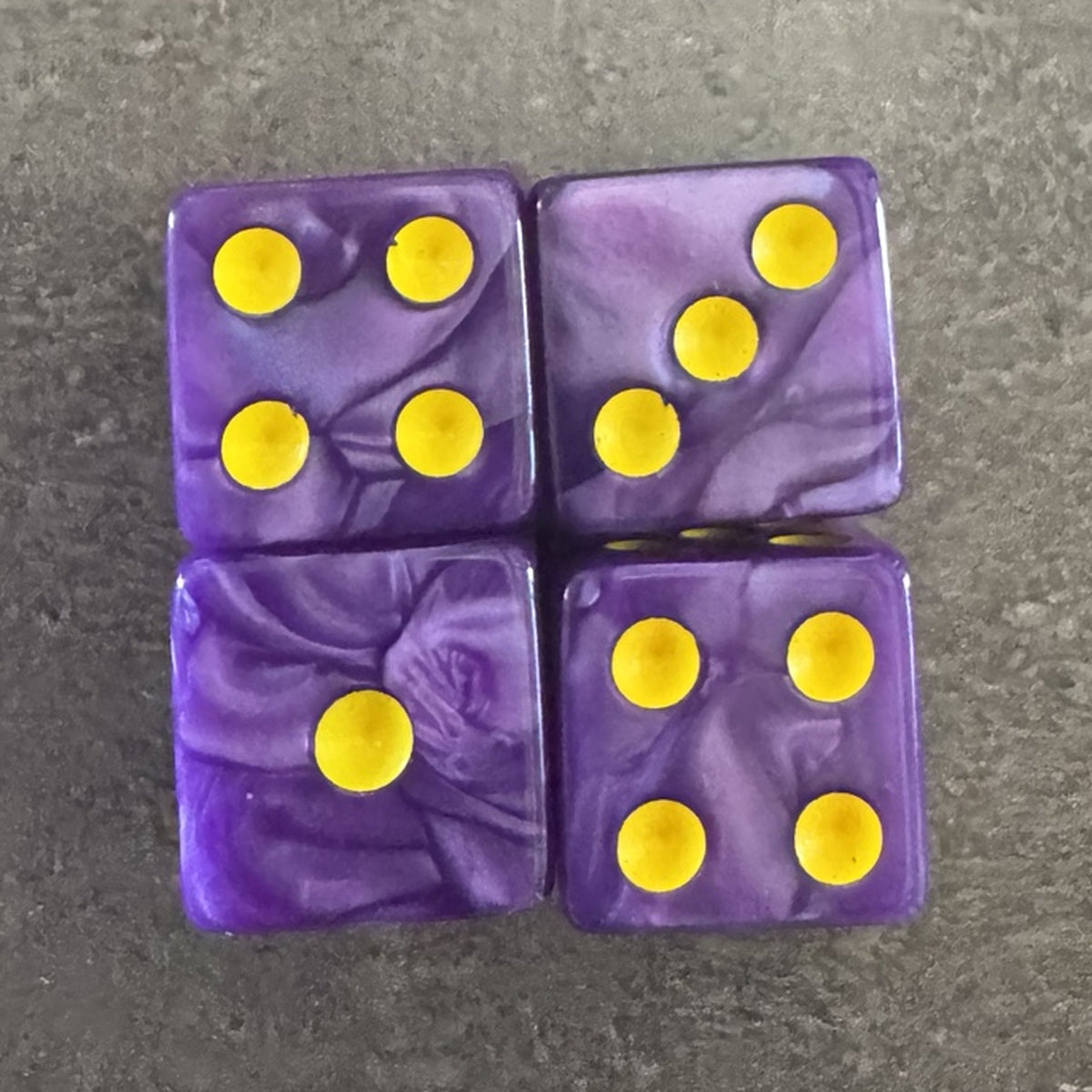 Photo réelle recadrée : 4 dés violets nacrés à points jaune doré. / Cropped original photo: 4 pearlescent purple D6 dice with yellow-gold pips.