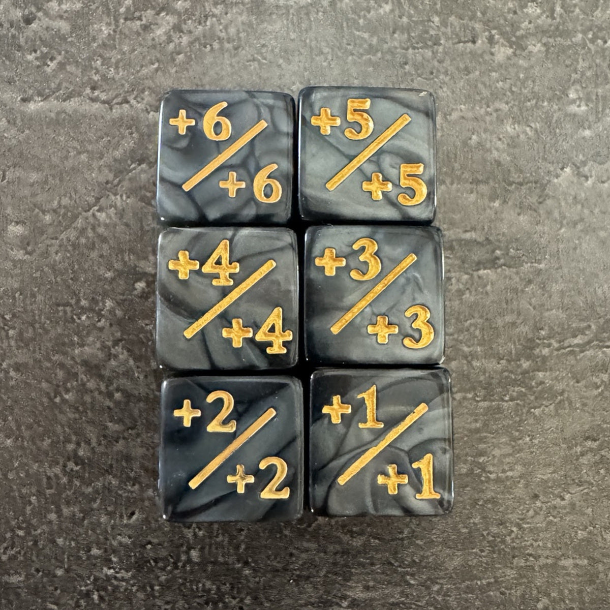 Lot de 6 dés compteurs positifs noirs et gris marbrés, marquages dorés +X/+X. / Set of 6 marbled black and grey positive counter dice with gold-coloured markings.
