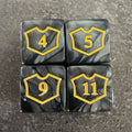 Photo réelle recadrée : lot de 4 dés compteurs noirs et gris, blasons dorés. / Cropped original photo: set of 4 black and grey counter dice with gold-coloured shield markings.