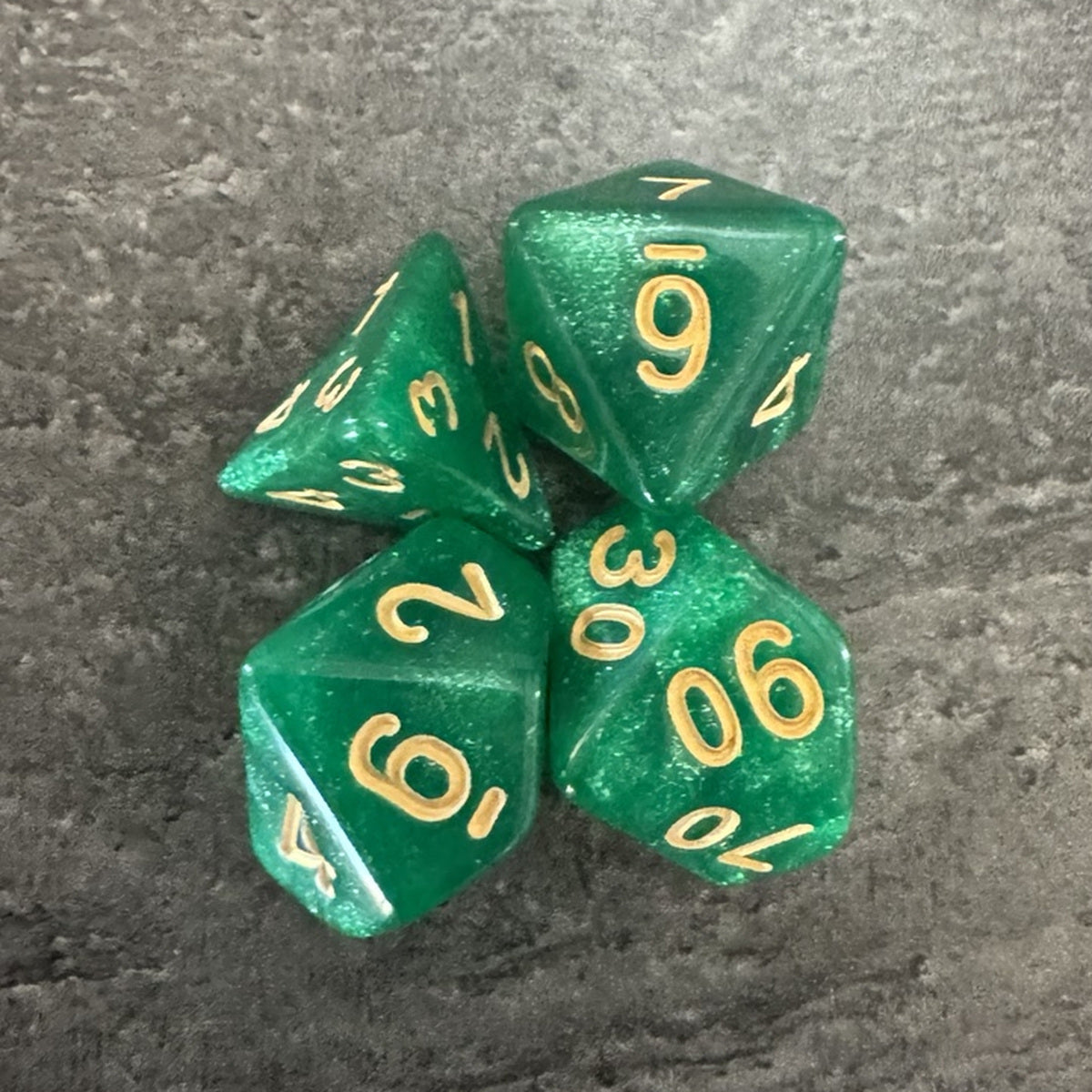Lot de 4 dés polyédriques verts pailletés, chiffres dorés. Photo réelle recadrée. / Assortment of 4 green glitter polyhedral dice with gold-coloured numbers. Cropped original photo.