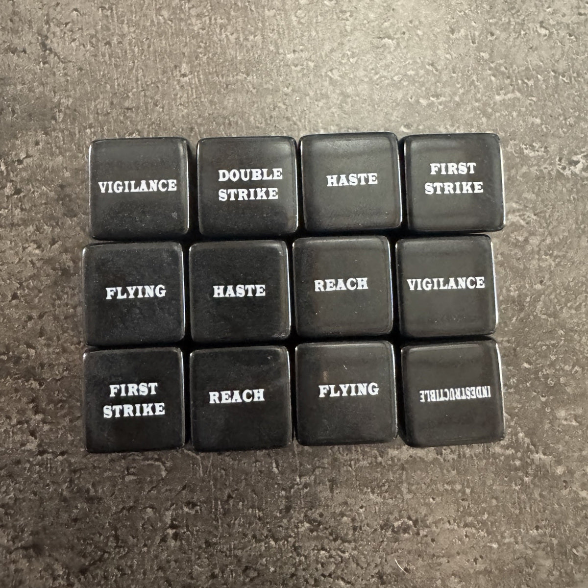 Lot de 12 dés noirs de capacités avec inscriptions blanches en anglais. Photo réelle recadrée. / Set of 12 black keyword dice with white English text. Cropped original photo.