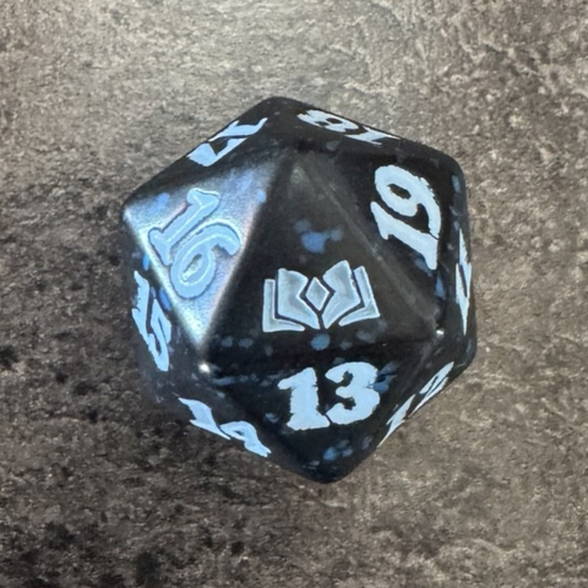 Dé D20 Magic Les Friches d'Eldraine, noir moucheté bleu à chiffres bleu clair — photo réelle. / Wilds of Eldraine blue Spindown D20 — actual photo.